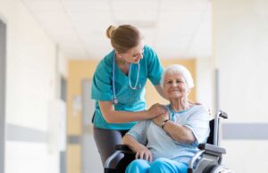 Older adults in a sunny common room doing gentle chair exercises with a facilitator nearby.